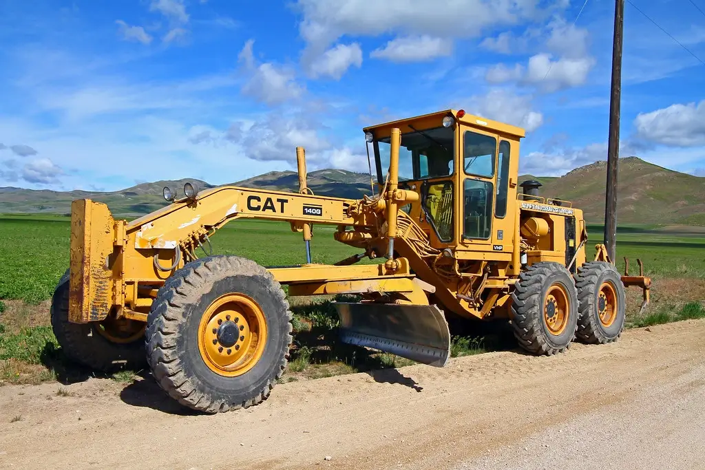 Yellow CAT 140G motor grader on a dirt road with green fields and hills.